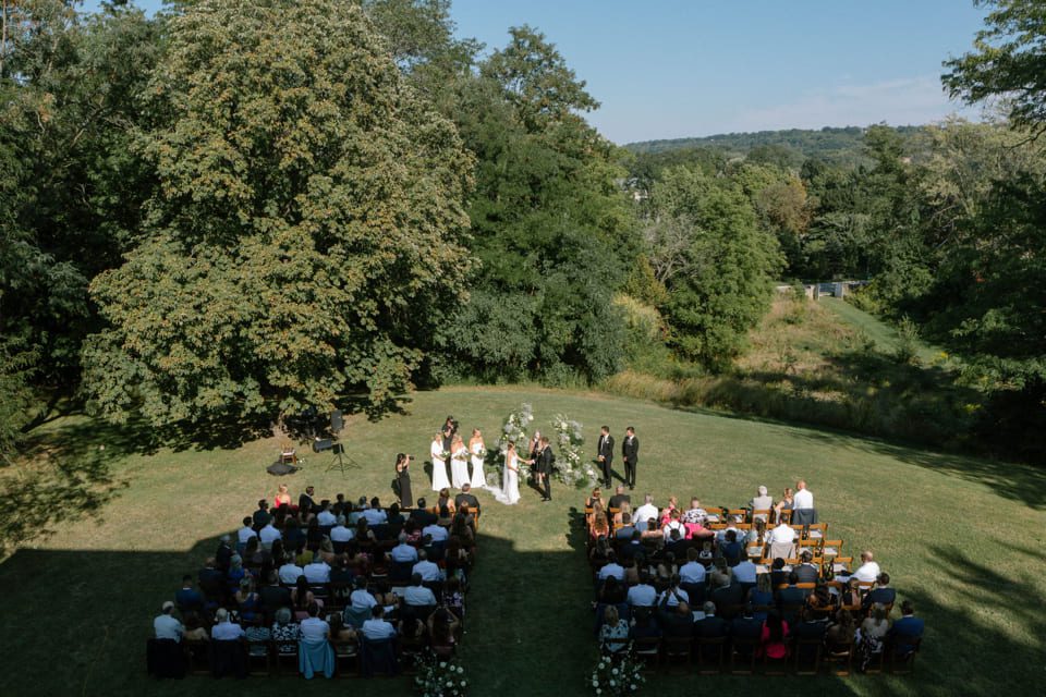 Aerial view of an outdoor wedding ceremony. Guests are seated facing a couple and officiant under a floral arch. The setting is grassy with large trees and a backdrop of hills under a clear blue sky. 124 on Queen Hotel and Spa | Hotel & Spa Niagara-on-the-lake | 1 (855) 988-4552