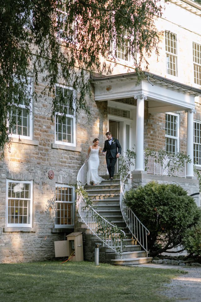 A bride in a white dress and groom in a dark suit descend stone steps of a historic building with a large porch and white columns. The building is covered in ivy, and sunlight filters through nearby trees. 124 on Queen Hotel and Spa | Hotel & Spa Niagara-on-the-lake | 1 (855) 988-4552