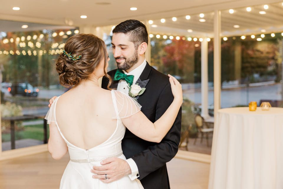 A bride and groom share a dance in a brightly lit room with large windows. The bride wears a white dress with delicate sleeves, and the groom is in a black suit with a green bow tie. The background shows string lights and a view of trees outside. 124 on Queen Hotel and Spa | Hotel & Spa Niagara-on-the-lake | 1 (855) 988-4552