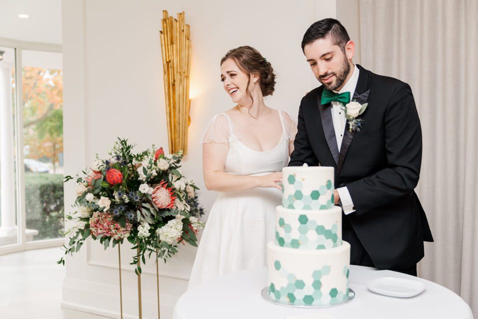 A bride and groom stand beside a three-tiered wedding cake decorated with green hexagons. The bride is smiling and wearing a white dress, while the groom, in a black suit, adjusts the cake. A floral arrangement is visible in the background. 124 on Queen Hotel and Spa | Hotel & Spa Niagara-on-the-lake | 1 (855) 988-4552