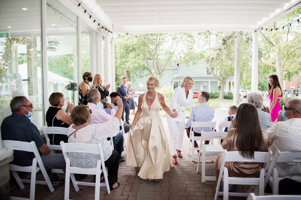A joyful wedding ceremony with two brides walking down the aisle together. One bride wears a white suit and the other a satin gown. Guests, some wearing masks, are seated on white chairs, applauding. The venue is a bright, covered outdoor space. 124 on Queen Hotel and Spa | Hotel & Spa Niagara-on-the-lake | 1 (855) 988-4552