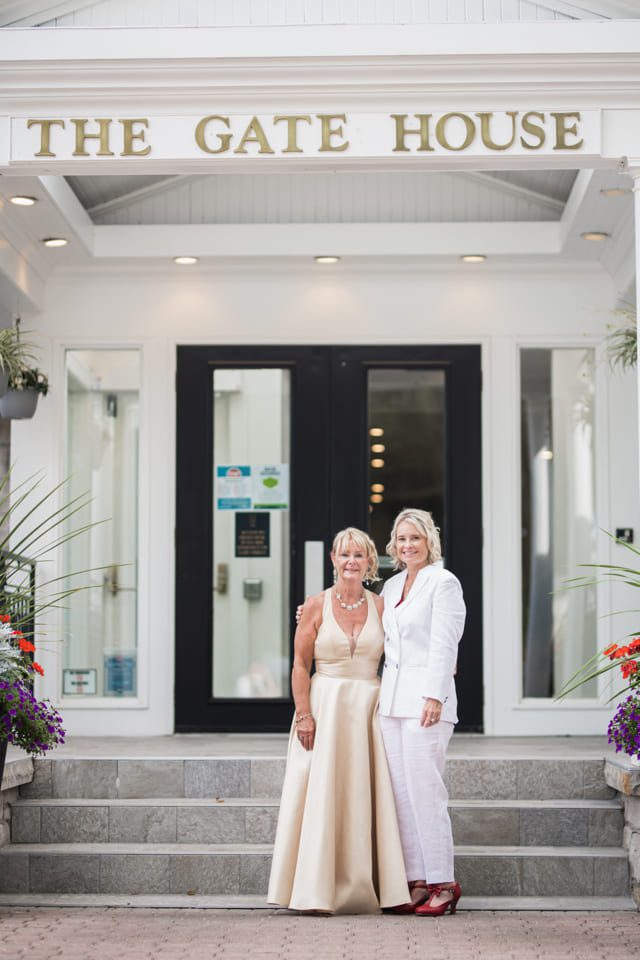 Two women are standing together in front of a building labeled The Gate House. One is wearing a light gown and the other a white suit with a white shirt. They are smiling, with colorful flowers visible on either side of the entrance. 124 on Queen Hotel and Spa | Hotel & Spa Niagara-on-the-lake | 1 (855) 988-4552