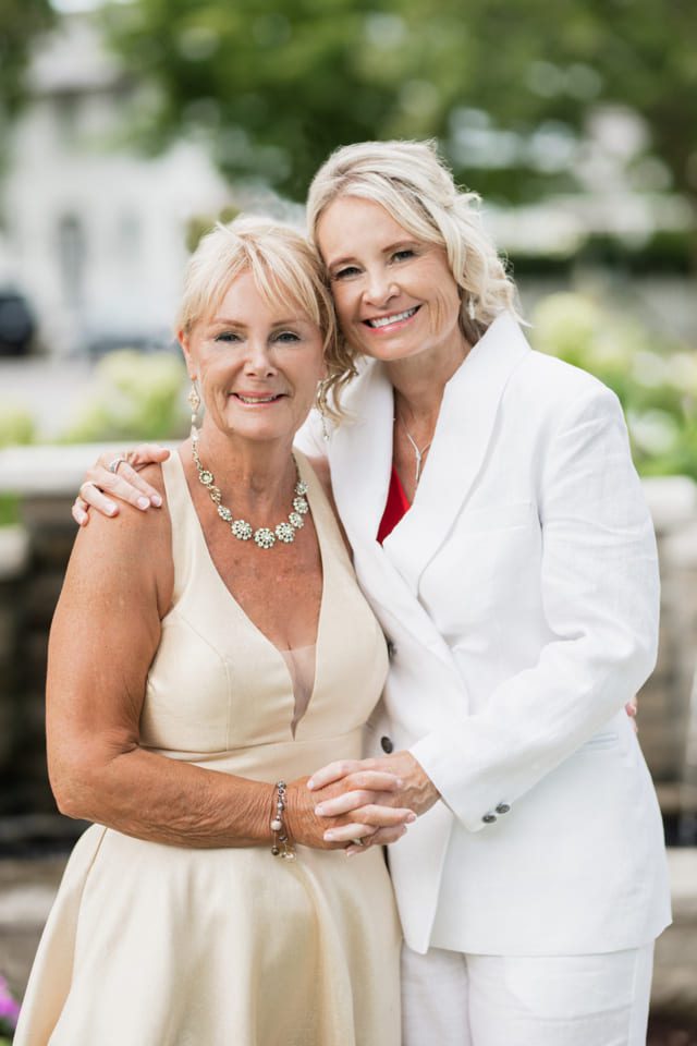 Two women smiling and embracing outdoors. One wears a beige dress with a floral necklace, the other a white suit with a red top. They stand in front of a blurred green and stone background, holding hands and looking at the camera. 124 on Queen Hotel and Spa | Hotel & Spa Niagara-on-the-lake | 1 (855) 988-4552