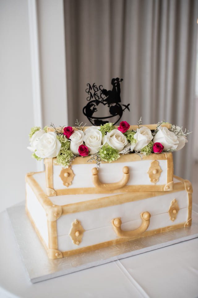 A wedding cake designed to resemble stacked vintage suitcases, decorated with white and pink roses on top. Theres a silhouette cake topper featuring a couple with the words Mr & Mrs. The setting is indoors with soft curtains in the background. 124 on Queen Hotel and Spa | Hotel & Spa Niagara-on-the-lake | 1 (855) 988-4552