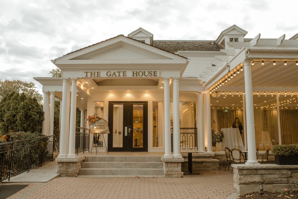 Front view of a white building labeled The Gate House with a porch and columns. Warm lights hang under the awning, and theres a small ramp and stairs leading to the entrance. Bushes are visible on the left, and the sky is overcast. 124 on Queen Hotel and Spa | Hotel & Spa Niagara-on-the-lake | 1 (855) 988-4552