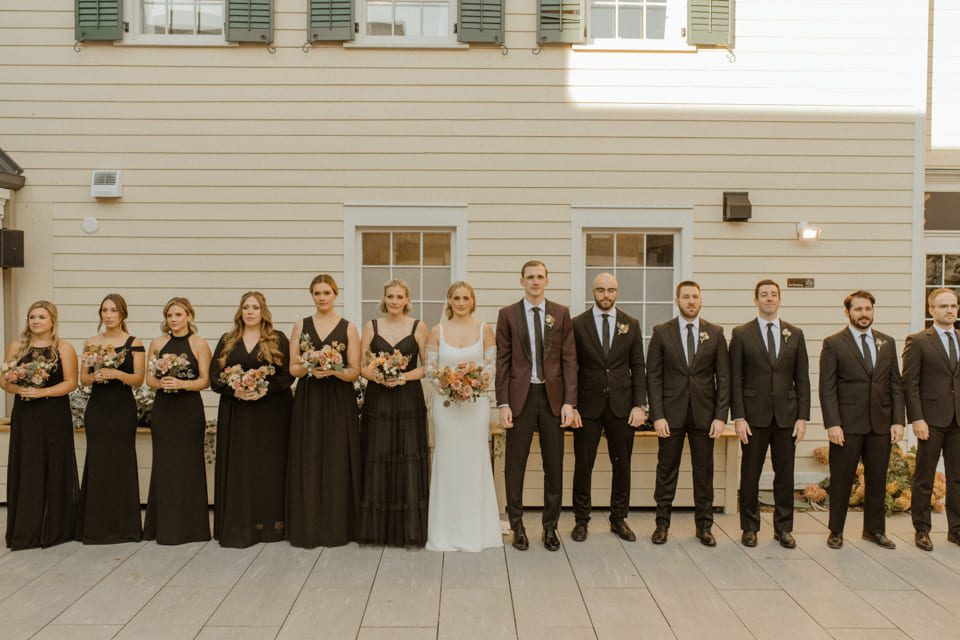 A wedding party stands in a line against a light-colored building. The group includes bridesmaids in black dresses and groomsmen in dark suits. The bride is in a white dress, holding a bouquet, and the groom is in a dark suit. 124 on Queen Hotel and Spa | Hotel & Spa Niagara-on-the-lake | 1 (855) 988-4552