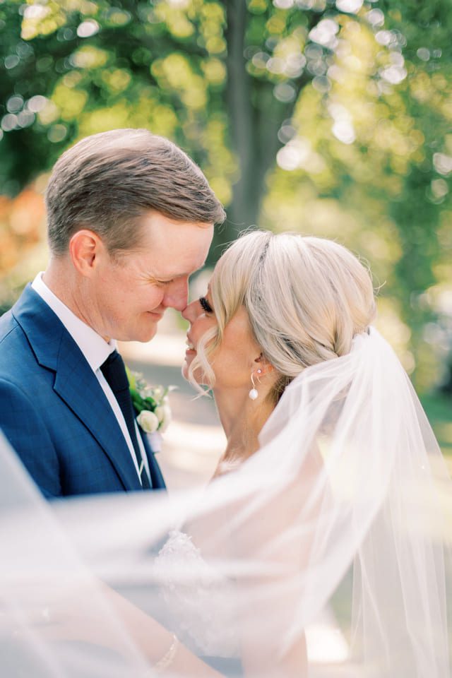A bride and groom share an intimate moment outdoors. The bride, in a white dress and veil, gazes lovingly at the groom, who is wearing a blue suit and tie. Sunlight filters through the trees in the background, creating a soft, romantic atmosphere. 124 on Queen Hotel and Spa | Hotel & Spa Niagara-on-the-lake | 1 (855) 988-4552