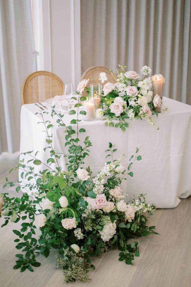 A beautifully arranged wedding table with a white tablecloth, adorned with light pink roses, greenery, and candles. A lush floral arrangement is placed in front of the table. Two wicker chairs are visible in the background. 124 on Queen Hotel and Spa | Hotel & Spa Niagara-on-the-lake | 1 (855) 988-4552