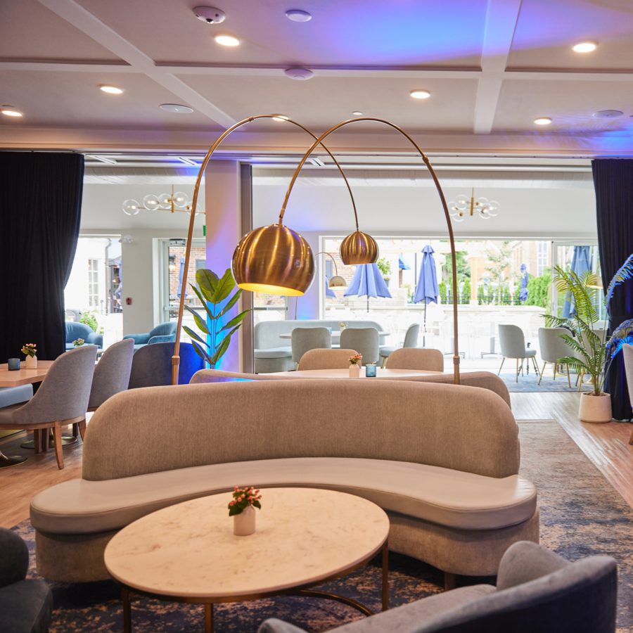 Luxurious lounge with modern decor, featuring a curved gray sofa, round marble table, and elegant gold floor lamp. Large windows reveal a patio with blue umbrellas. Plush chairs and plants enhance the spaces comfort and style. 124 on Queen Hotel and Spa | Hotel & Spa Niagara-on-the-lake | 1 (855) 988-4552