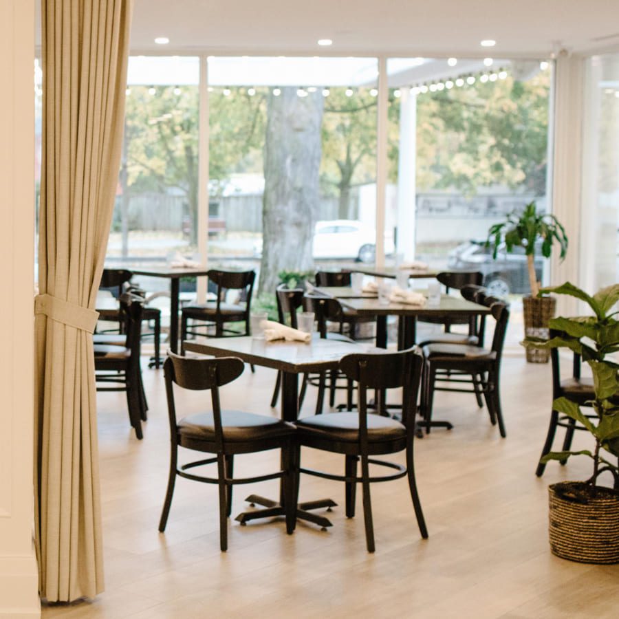 A bright, modern dining area with wooden floors, black chairs, and tables neatly set with napkins. Large windows reveal a view of trees outside. A potted plant and beige curtains add a touch of warmth to the inviting space. 124 on Queen Hotel and Spa | Hotel & Spa Niagara-on-the-lake | 1 (855) 988-4552