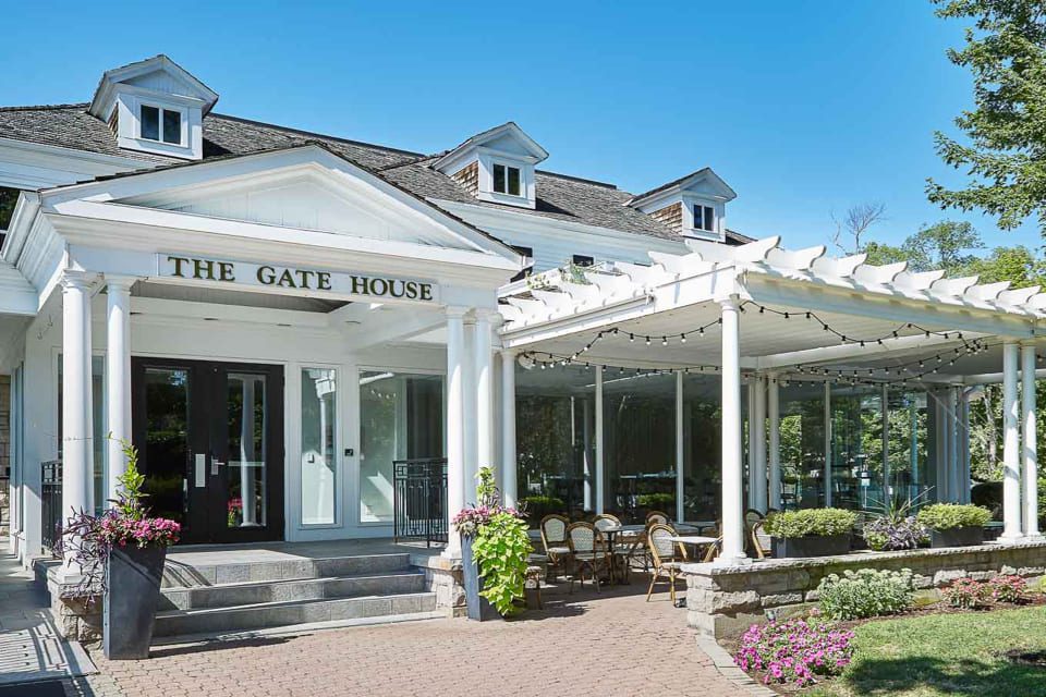 Historic white building with columns, labeled The Gate House. Features a covered terrace with string lights and patio seating. Surrounded by trees and flowers under a clear blue sky. 124 on Queen Hotel and Spa | Hotel & Spa Niagara-on-the-lake | 1 (855) 988-4552
