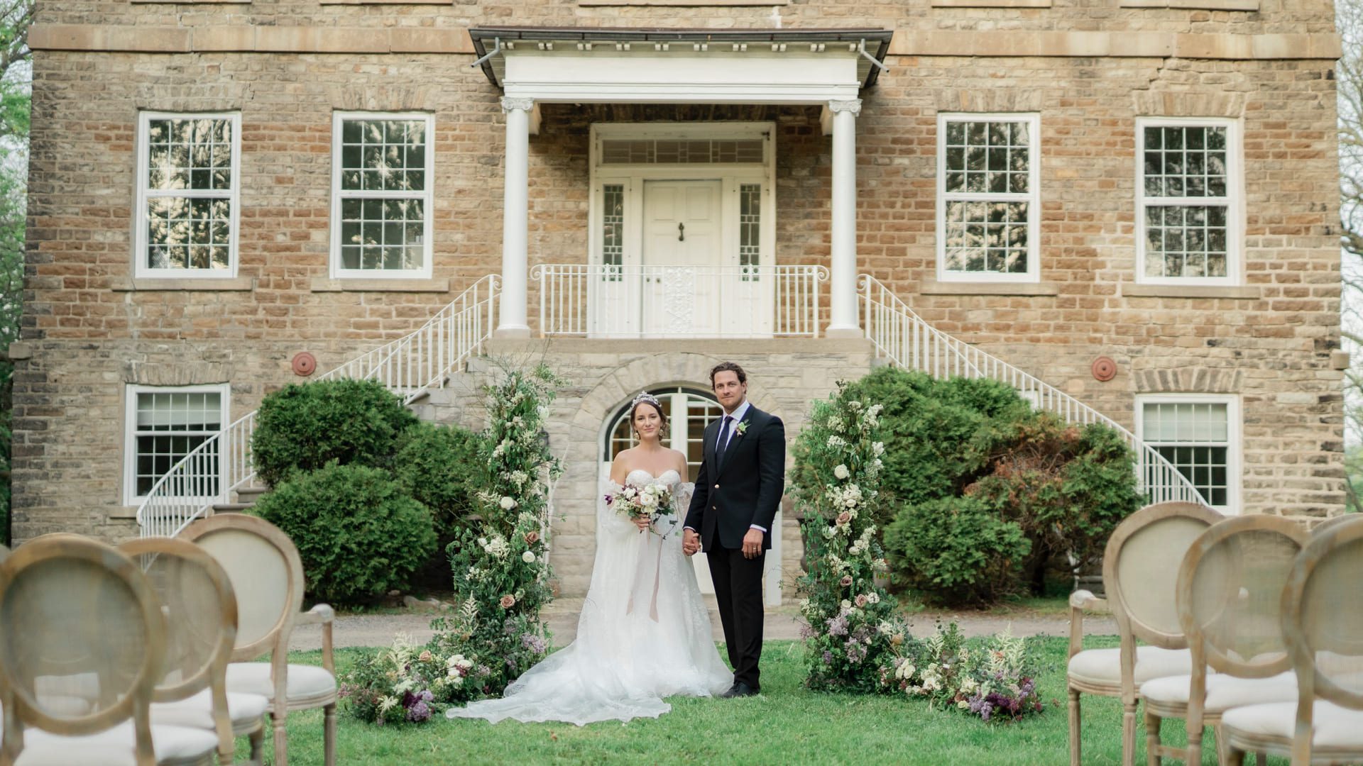 A bride and groom stand holding hands in front of a historic stone building with large windows and white doors. The couple is surrounded by lush greenery and flower arrangements. Empty chairs are arranged on the lawn in the foreground. 124 on Queen Hotel and Spa | Hotel & Spa Niagara-on-the-lake | 1 (855) 988-4552
