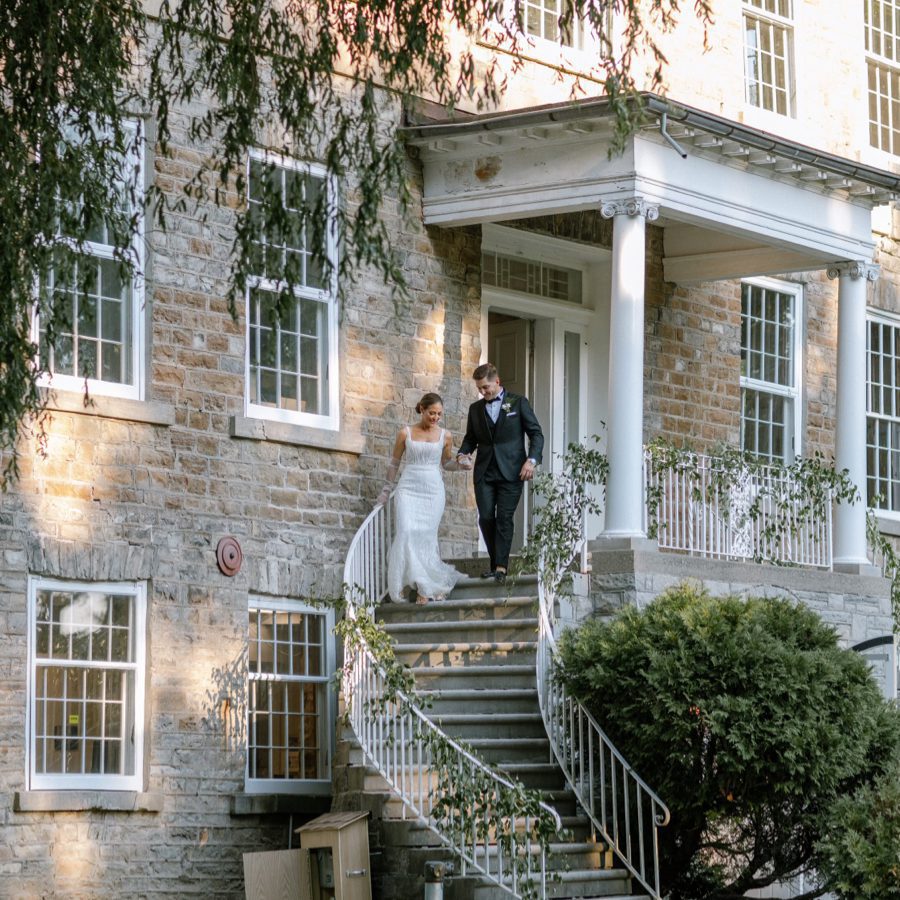 A bride and groom are walking down the steps of a large stone building with white columns and railings adorned with greenery. The bride is in a white dress, and the groom is in a black suit. The scene is set in a lush, green outdoor area. 124 on Queen Hotel and Spa | Hotel & Spa Niagara-on-the-lake | 1 (855) 988-4552