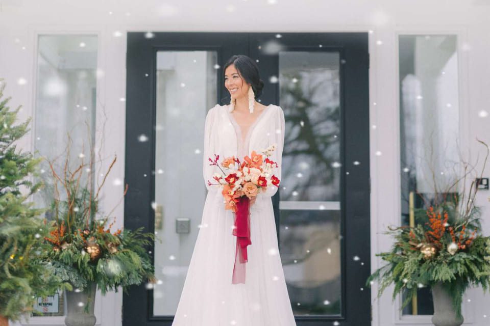 A bride in a white wedding dress stands outside in front of a dark door holding a vibrant bouquet of orange and red flowers. Snowflakes fall gently around her, and festive greenery with branches decorates each side of the entrance. 124 on Queen Hotel and Spa | Hotel & Spa Niagara-on-the-lake | 1 (855) 988-4552