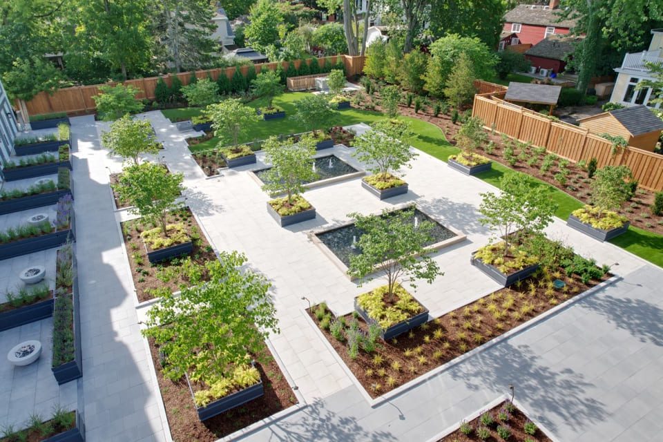 Aerial view of a modern, geometric garden with symmetrical pathways and rectangular planters filled with greenery and small trees. The garden is bordered by a wooden fence and surrounded by lush trees and neighboring houses. 124 on Queen Hotel and Spa | Hotel & Spa Niagara-on-the-lake | 1 (855) 988-4552 Aerial view of a modern, geometric garden with symmetrical pathways and rectangular planters filled with greenery and small trees. The garden is bordered by a wooden fence and surrounded by lush trees and neighboring houses. 124 on Queen Hotel and Spa | Hotel & Spa Niagara-on-the-lake | 1 (855) 988-4552