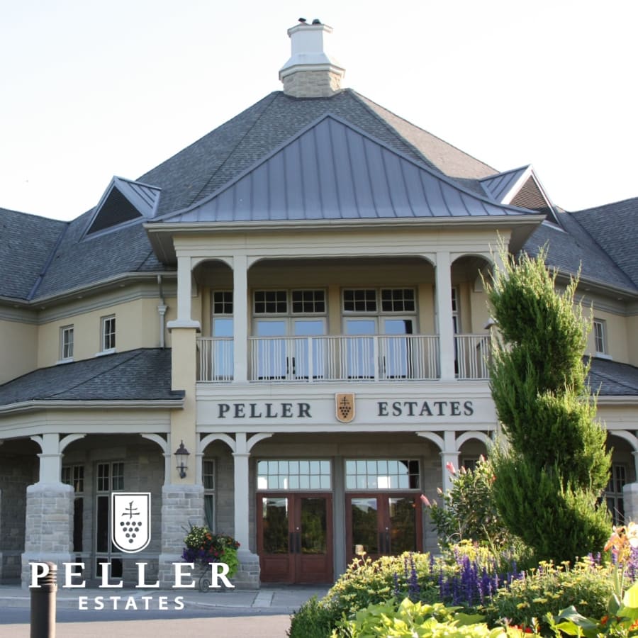 A large, elegant building with a peaked roof and tall columns, labeled Peller Estates above the entrance. The foreground features lush green foliage and flowers. The logo Peller Estates is visible in the bottom corner. 124 on Queen Hotel and Spa | Hotel & Spa Niagara-on-the-lake | 1 (855) 988-4552