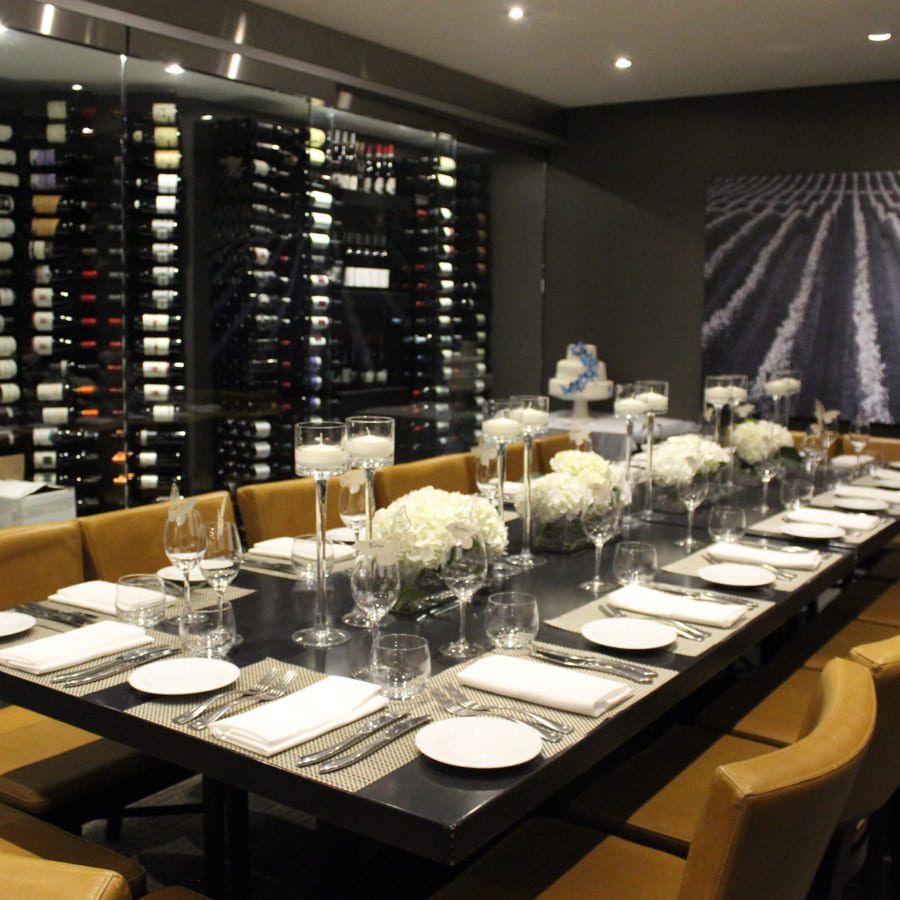 Elegant dining room with a long table set for a meal. White flowers and high candles decorate the center. Surrounding wine racks display various bottles. Brown cushioned chairs line the table, and a large photo of a vineyard hangs on the wall. 124 on Queen Hotel and Spa | Hotel & Spa Niagara-on-the-lake | 1 (855) 988-4552
