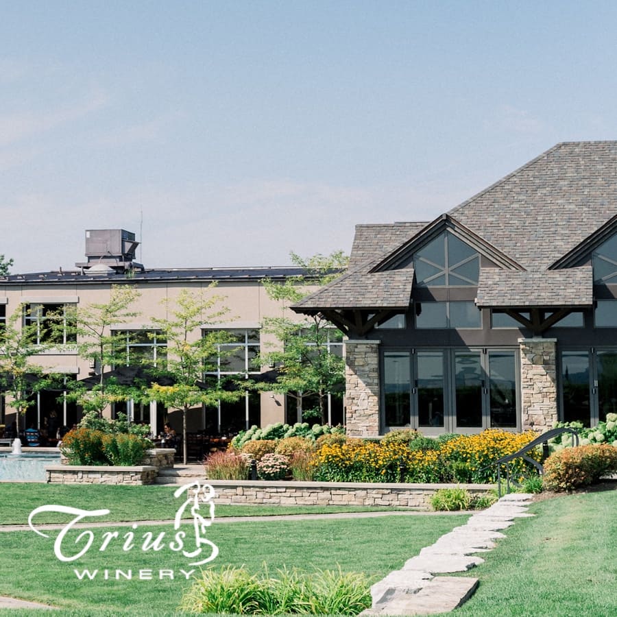 Charming winery building with large windows and stone accents surrounded by lush green lawn and colorful flower beds under a clear blue sky. The words Trius Winery are visible in the bottom left corner. 124 on Queen Hotel and Spa | Hotel & Spa Niagara-on-the-lake | 1 (855) 988-4552