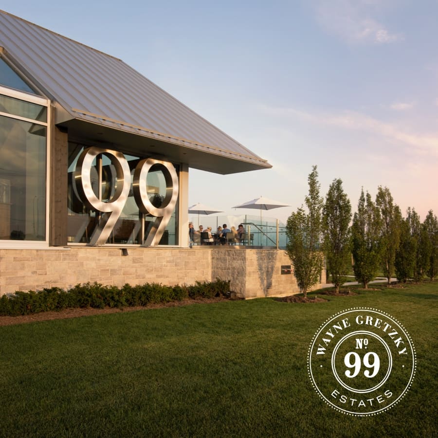 Modern building with large 99 on the side, surrounded by a neatly maintained lawn and trees. People are seated on a terrace under umbrellas. The bottom corner has a Wayne Gretzky Estates No. 99 logo. Sky is clear and blue. 124 on Queen Hotel and Spa | Hotel & Spa Niagara-on-the-lake | 1 (855) 988-4552