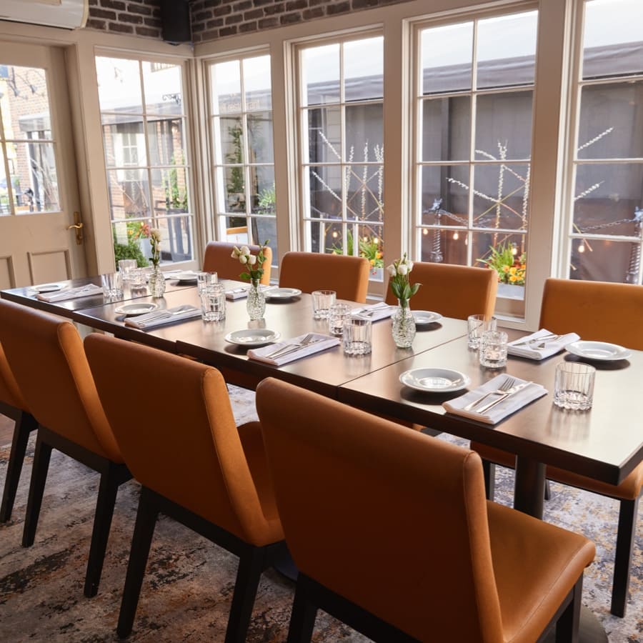 A sunlit dining room with a long wooden table set for eight. Orange chairs surround the table, which is adorned with glassware, plates, and small vases with flowers. Large windows allow natural light to fill the space, highlighting the brick walls. 124 on Queen Hotel and Spa | Hotel & Spa Niagara-on-the-lake | 1 (855) 988-4552