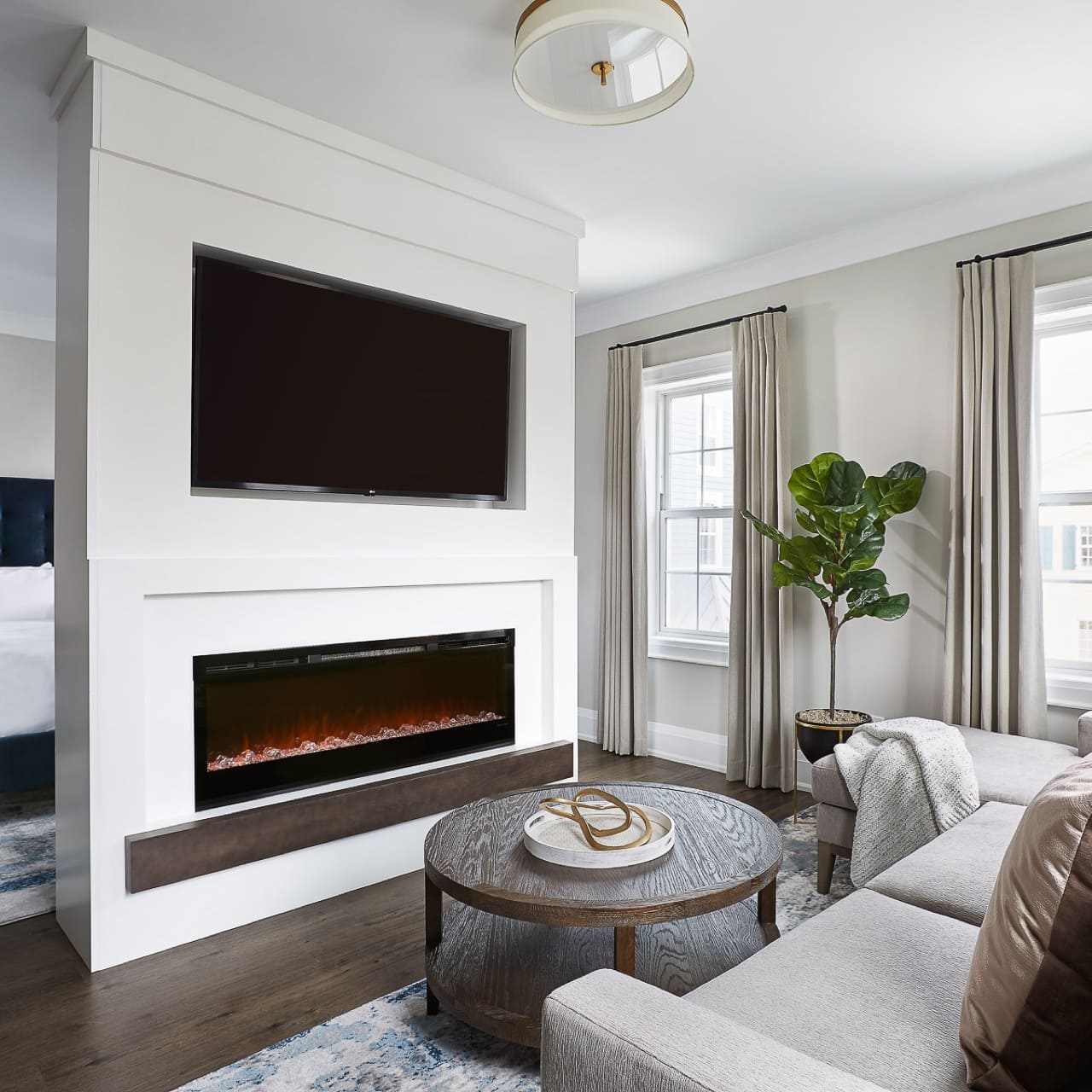 Modern living room with a large wall-mounted TV and an electric fireplace beneath it. A gray sectional sofa faces a round wooden coffee table. Large windows with beige curtains and a potted plant add to the bright, cozy atmosphere. 124 on Queen Hotel and Spa | Hotel & Spa Niagara-on-the-lake | 1 (855) 988-4552