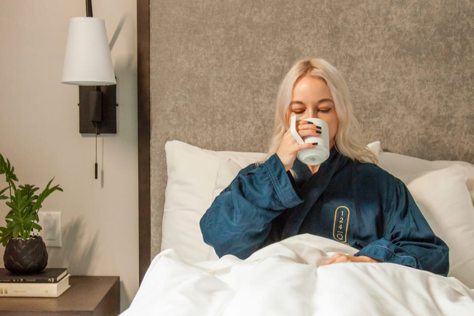 A person with long blonde hair is sitting in bed, wearing a blue robe, and drinking from a mug. They appear relaxed. A small plant and a book are on the bedside table. A wall lamp illuminates the scene. 124 on Queen Hotel and Spa | Hotel & Spa Niagara-on-the-lake | 1 (855) 988-4552