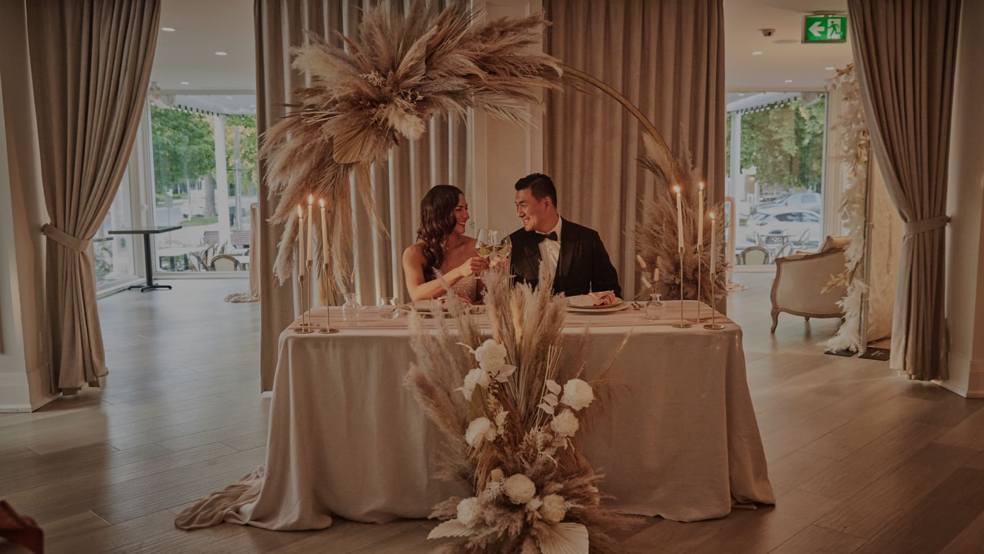 A bride and groom sit at a decorated table with an arch of pampas grass and flowers. They are toasting with champagne in a softly lit room, surrounded by elegant drapery and candles. Large windows reveal a view of trees outside. 124 on Queen Hotel and Spa | Hotel & Spa Niagara-on-the-lake | 1 (855) 988-4552