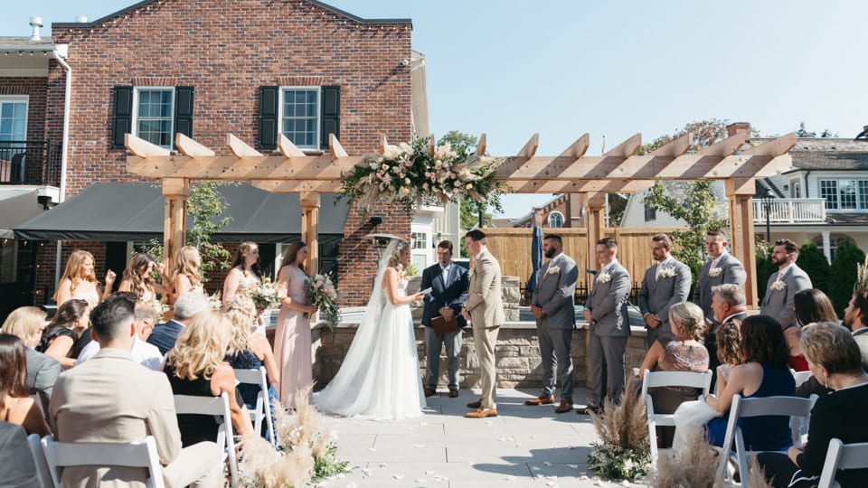 A bride and groom stand under a pergola during an outdoor wedding ceremony. They are surrounded by bridesmaids and groomsmen on a sunny day, with guests seated on either side. The backdrop includes a red brick building. 124 on Queen Hotel and Spa | Hotel & Spa Niagara-on-the-lake | 1 (855) 988-4552