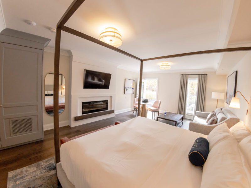 Luxurious hotel room with a four-poster bed, white bedding, armchair, and couch. A TV is mounted above a modern fireplace. A small dining table with chairs is near a large window, allowing natural light to brighten the room. Neutral color palette. 124 on Queen Hotel and Spa | Hotel & Spa Niagara-on-the-lake | 1 (855) 988-4552