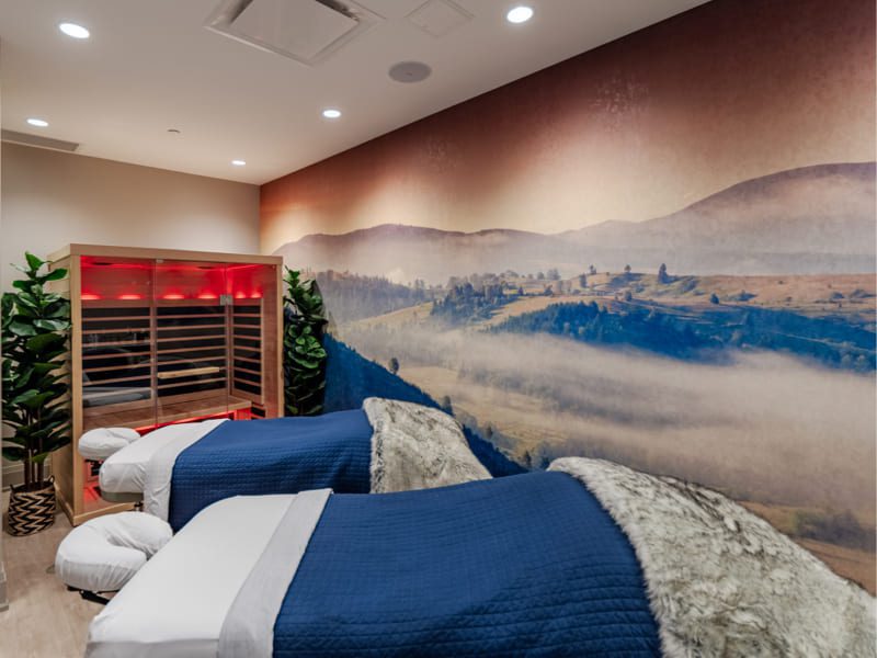 A serene spa room with two massage beds covered in blue blankets and fur throws. A large mural of a misty mountain landscape decorates the wall. A small infrared sauna glows with red light in the corner, surrounded by potted plants. 124 on Queen Hotel and Spa | Hotel & Spa Niagara-on-the-lake | 1 (855) 988-4552