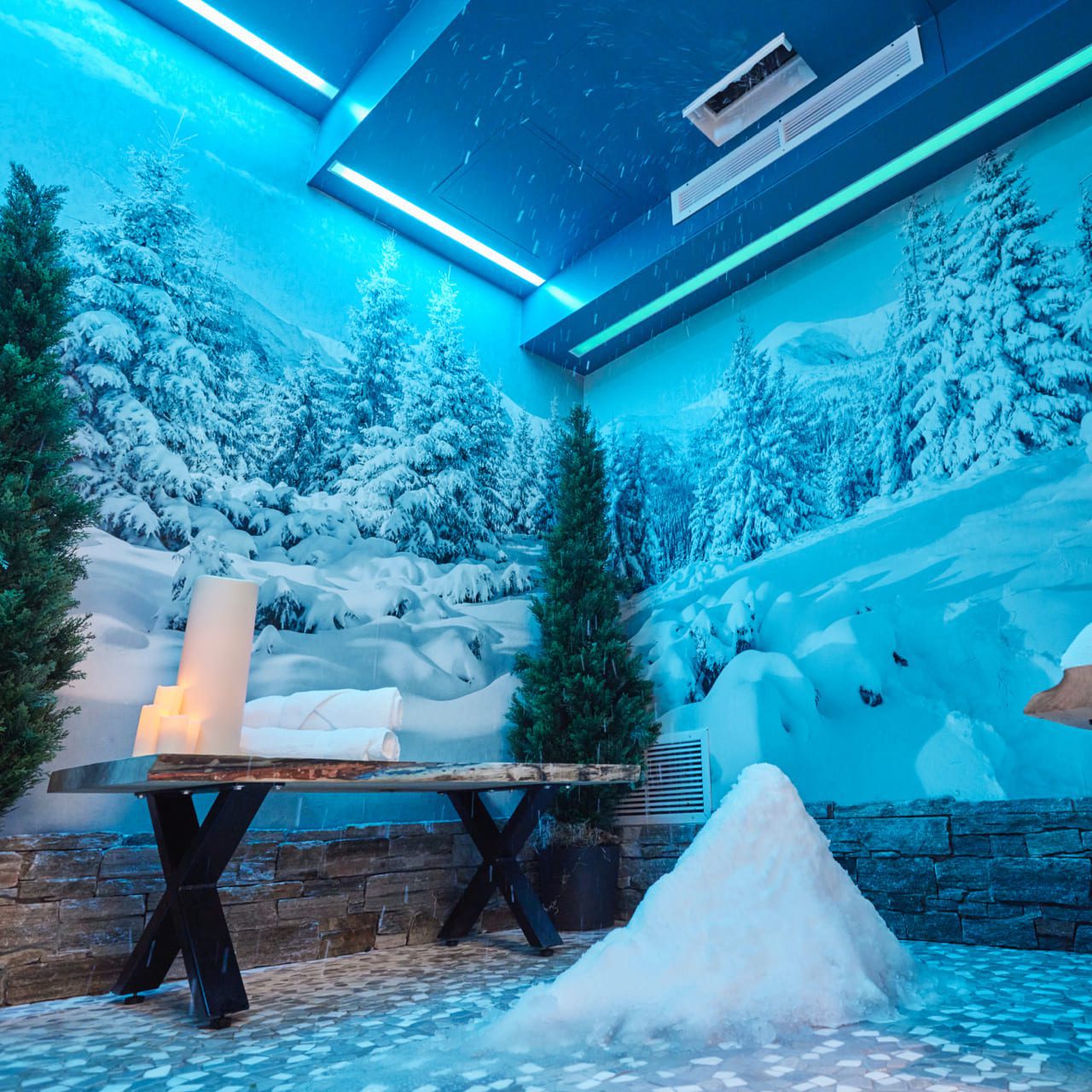 A room with blue lighting features a faux snowy pine forest mural. Theres a wooden table with candles, a small pile of fake snow on the floor, and two potted trees, creating a winter-themed atmosphere. 124 on Queen Hotel and Spa | Hotel & Spa Niagara-on-the-lake | 1 (855) 988-4552