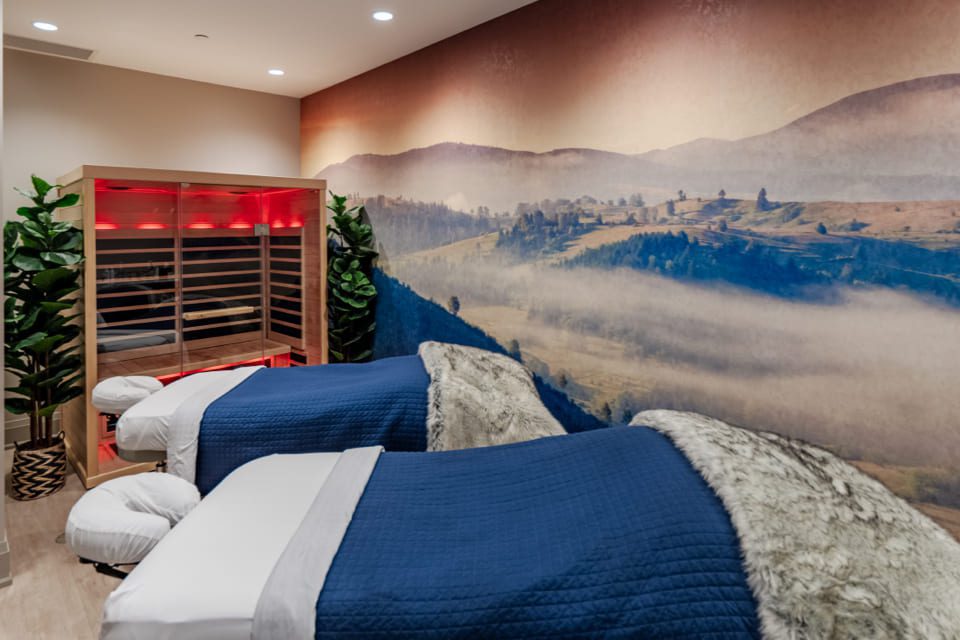 A spa room with two massage tables covered in blue blankets and fur throws. A mural of a misty mountain landscape decorates the wall. There are potted plants and an infrared sauna in the corner with a warm red glow. 124 on Queen Hotel and Spa | Hotel & Spa Niagara-on-the-lake | 1 (855) 988-4552