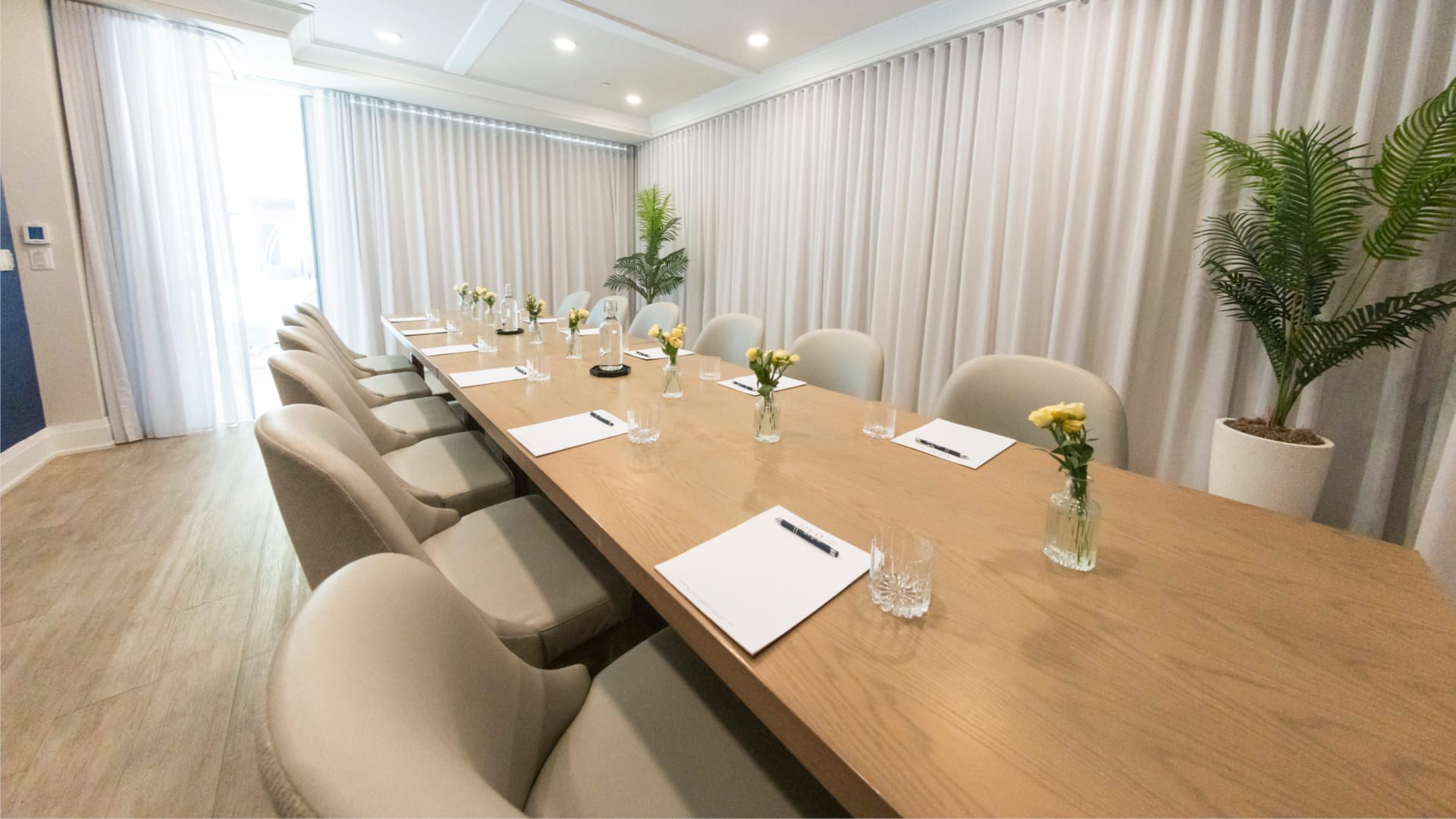 A modern conference room with a long wooden table surrounded by beige chairs. The table is set with notepads, pens, and small vases holding yellow flowers. Large windows with sheer curtains and potted plants enhance the rooms ambiance. 124 on Queen Hotel and Spa | Hotel & Spa Niagara-on-the-lake | 1 (855) 988-4552