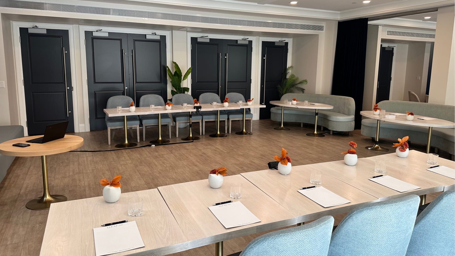 Conference room setup with a U-shaped arrangement of tables and chairs, each having a notepad and glass. Small vases with orange flowers decorate the tables. Black doors line the back wall, with potted plants and a beige curved bench on the right. 124 on Queen Hotel and Spa | Hotel & Spa Niagara-on-the-lake | 1 (855) 988-4552