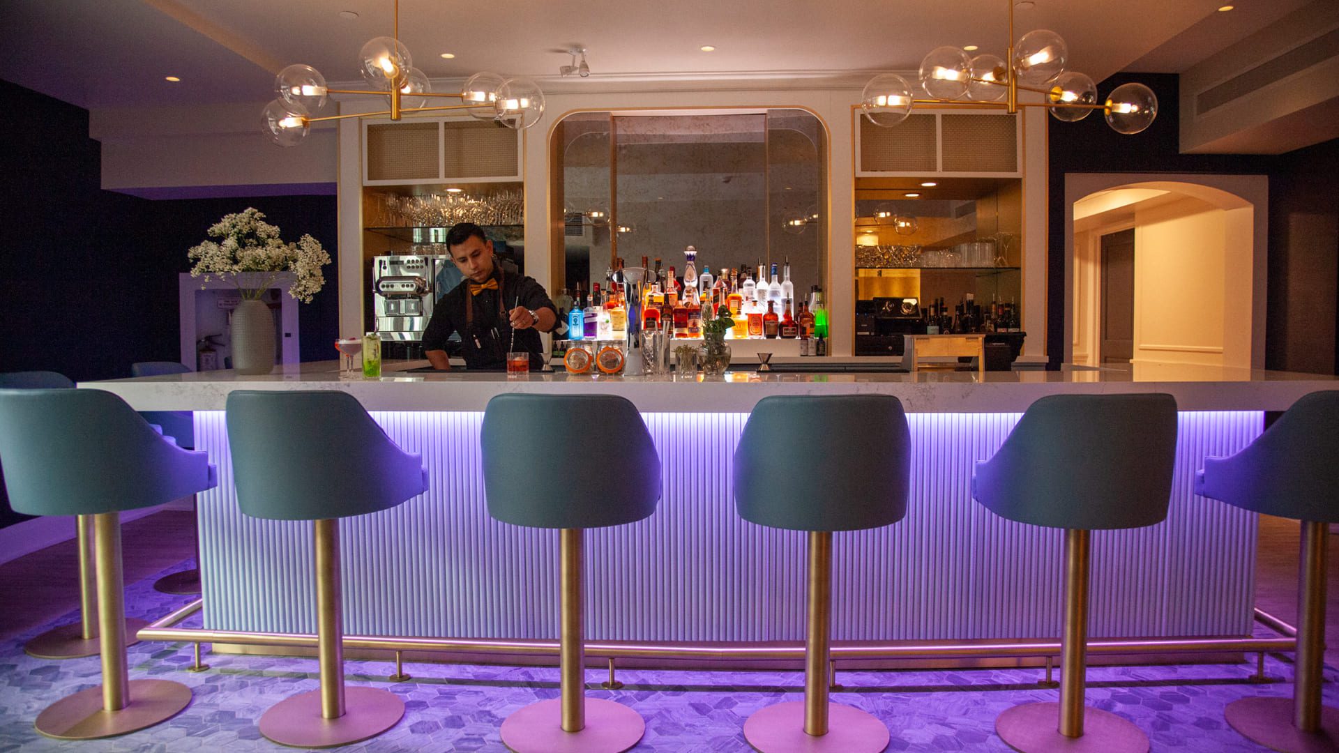 A modern bar with a sleek white counter and five high-backed chairs, subtly illuminated in purple. A bartender prepares drinks amid a backdrop of neatly arranged bottles and glasses. The setting is stylish with decorative lighting above. 124 on Queen Hotel and Spa | Hotel & Spa Niagara-on-the-lake | 1 (855) 988-4552
