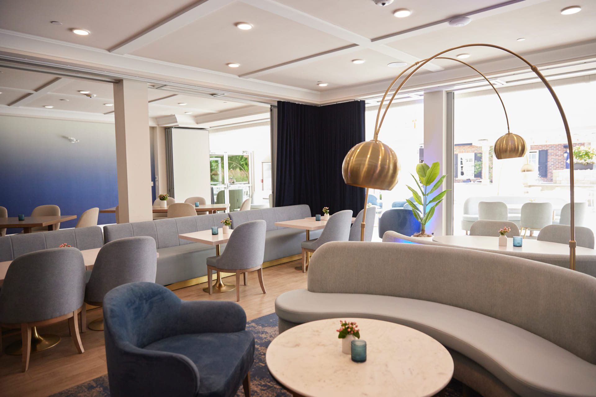 Modern cafe interior with gray and blue seating, round white marble tables, and a tall, curved brass floor lamp. Large windows allow natural light to fill the space. A small plant decorates each table. 124 on Queen Hotel and Spa | Hotel & Spa Niagara-on-the-lake | 1 (855) 988-4552