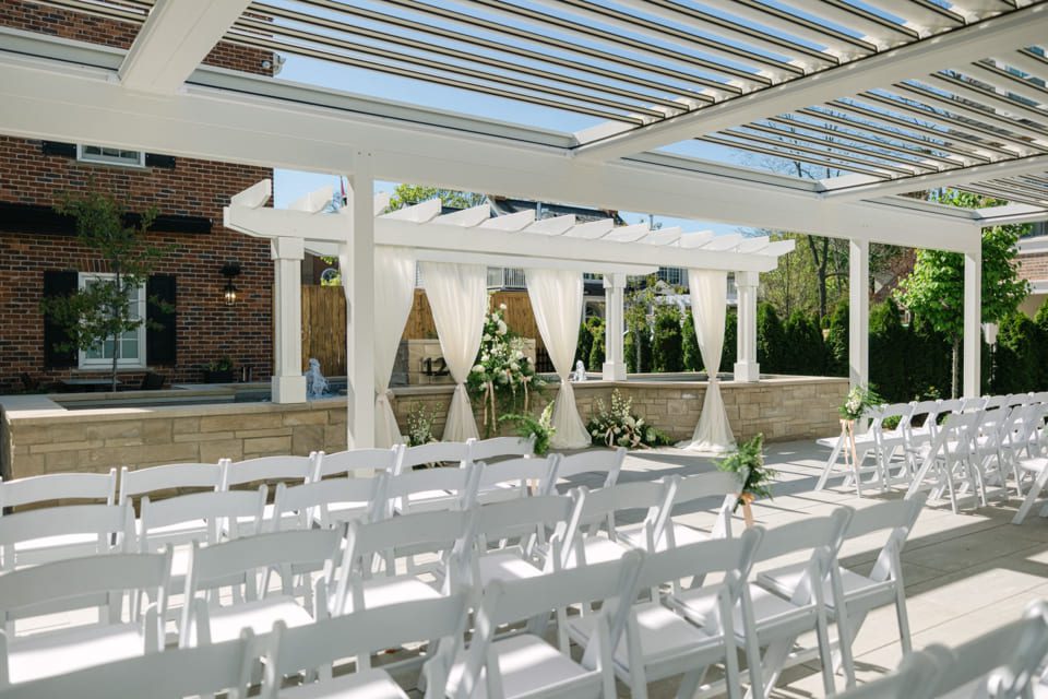 Outdoor wedding venue with rows of white chairs facing a decorated arbor with white drapes and floral arrangements. Sunny, clear skies enhance the garden-like setting with a brick building in the background. 124 on Queen Hotel and Spa | Hotel & Spa Niagara-on-the-lake | 1 (855) 988-4552 Outdoor wedding venue with rows of white chairs facing a decorated arbor with white drapes and floral arrangements. Sunny, clear skies enhance the garden-like setting with a brick building in the background. 124 on Queen Hotel and Spa | Hotel & Spa Niagara-on-the-lake | 1 (855) 988-4552
