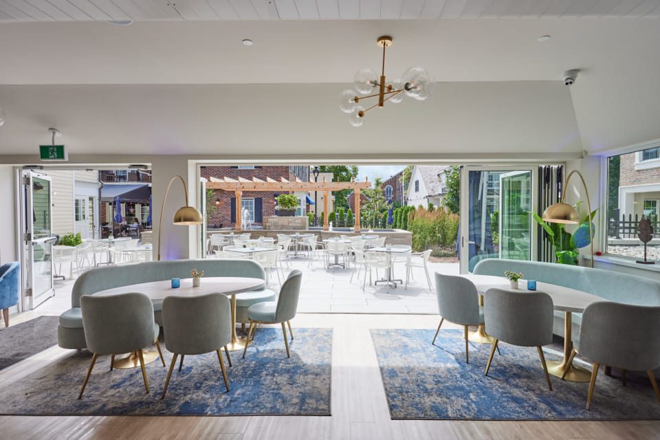 Modern restaurant interior with light blue seating, round white tables, and brass accents. Large windows open to an outdoor patio with more seating. Natural light pours in, highlighting the contemporary design and spacious layout. 124 on Queen Hotel and Spa | Hotel & Spa Niagara-on-the-lake | 1 (855) 988-4552