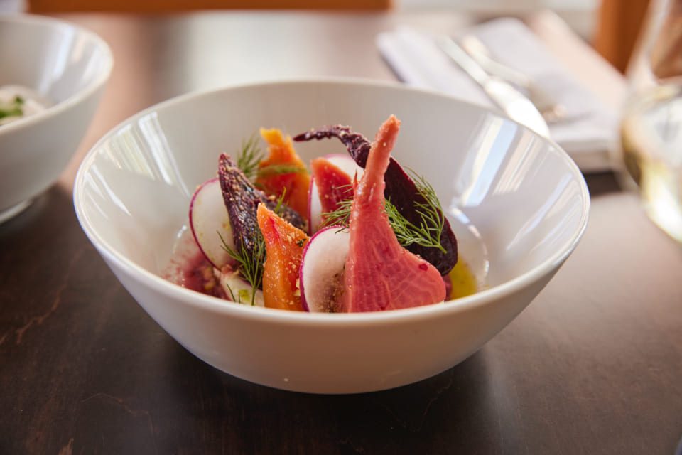 A white bowl contains a colorful salad with sliced red and orange beets, radishes, and garnished with fresh dill. The bowl is placed on a dark wooden table next to a pair of cutlery wrapped in a napkin. 124 on Queen Hotel and Spa | Hotel & Spa Niagara-on-the-lake | 1 (855) 988-4552