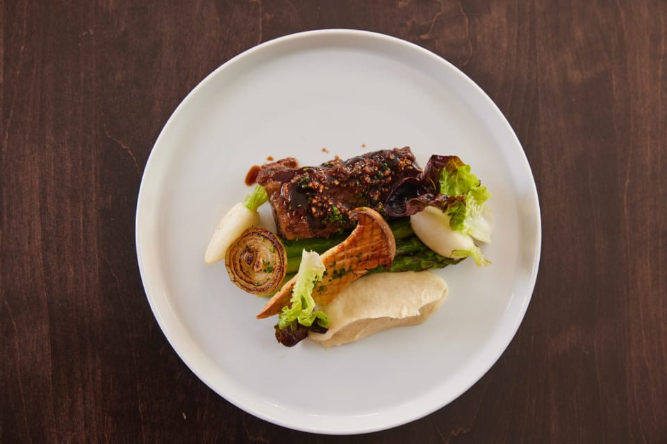 A gourmet dish on a white plate featuring glazed meat, asparagus, roasted onion, greens, and a dollop of creamy puree, artfully arranged on a dark wooden table. 124 on Queen Hotel and Spa | Hotel & Spa Niagara-on-the-lake | 1 (855) 988-4552
