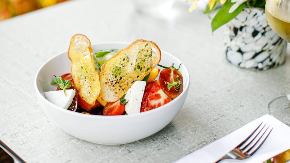 A white bowl brimming with a vibrant salad of assorted tomatoes, mozzarella slices, and fresh greens, topped with crispy bread slices, captures the essence of dining at one of the best 5 must-try restaurants in Niagara-on-the-Lake. Its set on a textured table beside a plant and cutlery. 124 on Queen Hotel and Spa | Hotel & Spa Niagara-on-the-lake | 1 (855) 988-4552