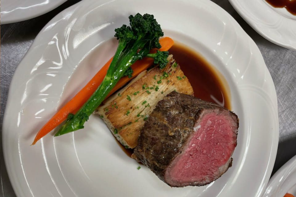 A gourmet dish featuring a medium-rare beef fillet with a rich brown sauce, garnished with sliced green herbs. Accompanied by a piece of crispy potato terrine, a cooked carrot, and a stalk of broccoli on a white plate. 124 on Queen Hotel and Spa | Hotel & Spa Niagara-on-the-lake | 1 (855) 988-4552