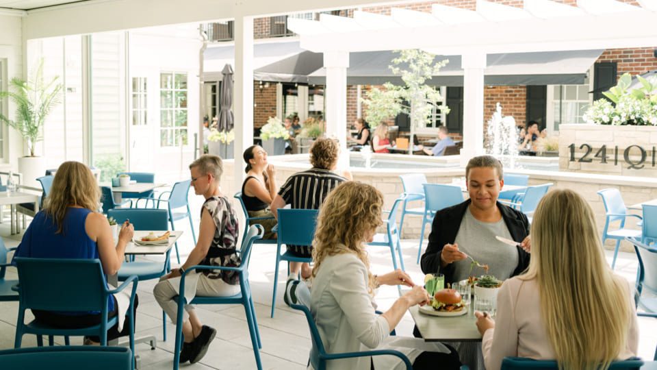 People are dining in an outdoor restaurant patio, exploring the newly enhanced patio at NOTL Bar & Restaurant. Several individuals are seated at blue tables, enjoying meals. A fountain and more diners can be seen in the background under shaded areas. The atmosphere is relaxed and casual. 124 on Queen Hotel and Spa | Hotel & Spa Niagara-on-the-lake | 1 (855) 988-4552