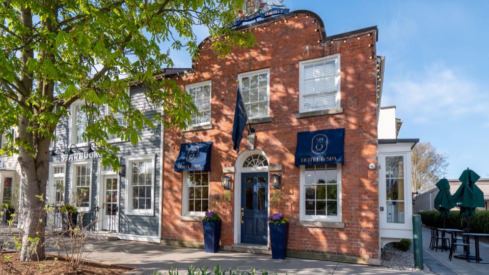 A charming brick building with a blue door, framed by trees, houses the Hotel & Spa. Next to it, explore the newly enhanced patio at NOTL Bar & Restaurant with green umbrellas. Its a bright, sunny day under a clear blue sky. 124 on Queen Hotel and Spa | Hotel & Spa Niagara-on-the-lake | 1 (855) 988-4552