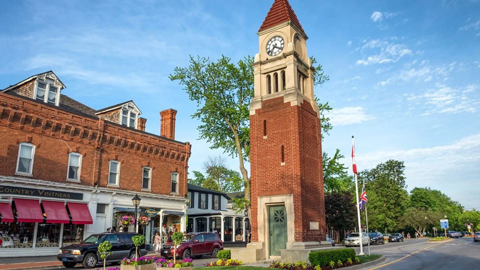 A red brick clock tower stands in a charming town square, surrounded by historic buildings. Nearby, explore the newly enhanced patio at NOTL Bar & Restaurant. A shop with a red awning is on the left, and a tree adds a touch of nature. A clear blue sky completes the picturesque scene. 124 on Queen Hotel and Spa | Hotel & Spa Niagara-on-the-lake | 1 (855) 988-4552