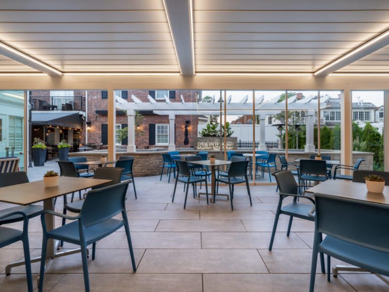 Outdoor patio with blue chairs and tables under a white pergola. The area is well-lit with neon lights on the ceiling. Small potted plants adorn some tables. A brick building and greenery are visible in the background. 124 on Queen Hotel and Spa | Hotel & Spa Niagara-on-the-lake | 1 (855) 988-4552