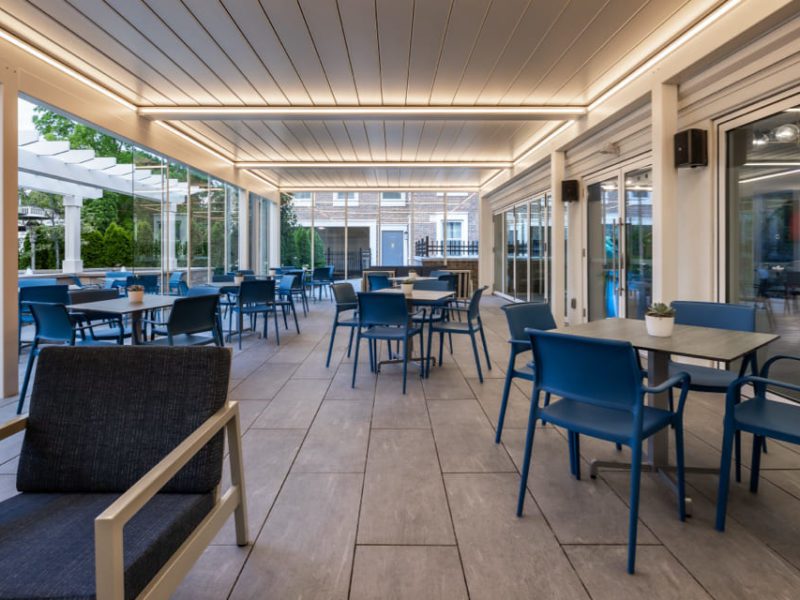 Outdoor dining area with modern decor, featuring blue chairs and square tables. The space is covered with a lighted pergola and surrounded by glass sliding doors and greenery. The tiled floor complements the clean, contemporary design. 124 on Queen Hotel and Spa | Hotel & Spa Niagara-on-the-lake | 1 (855) 988-4552