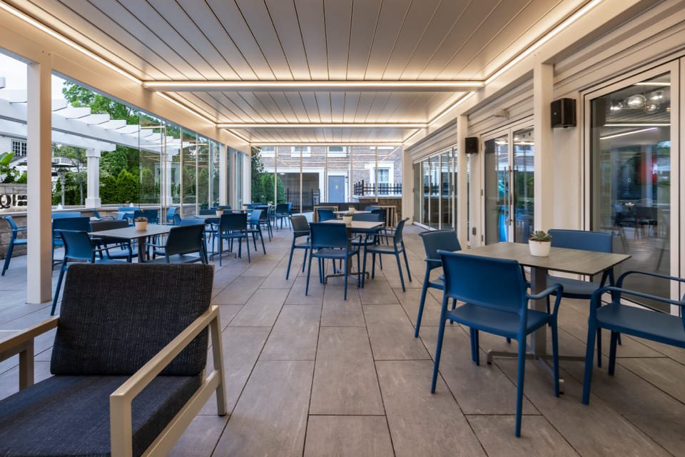 Outdoor dining area with modern decor, featuring blue chairs and square tables. The space is covered with a lighted pergola and surrounded by glass sliding doors and greenery. The tiled floor complements the clean, contemporary design. 124 on Queen Hotel and Spa | Hotel & Spa Niagara-on-the-lake | 1 (855) 988-4552
