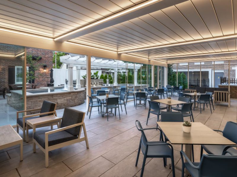 Outdoor patio with modern seating and tables. Glass walls provide a view of a brick building and surrounding greenery. Overhead, a slatted roof with integrated lighting adds an ambient atmosphere. Some tables have small potted plants. 124 on Queen Hotel and Spa | Hotel & Spa Niagara-on-the-lake | 1 (855) 988-4552