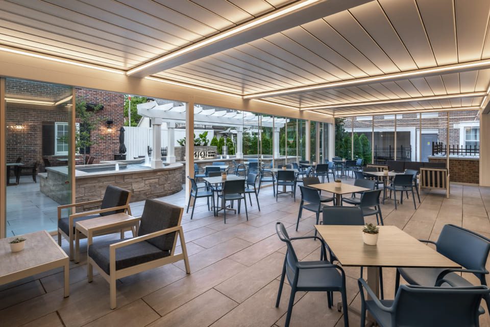 Outdoor patio with modern seating and tables. Glass walls provide a view of a brick building and surrounding greenery. Overhead, a slatted roof with integrated lighting adds an ambient atmosphere. Some tables have small potted plants. 124 on Queen Hotel and Spa | Hotel & Spa Niagara-on-the-lake | 1 (855) 988-4552